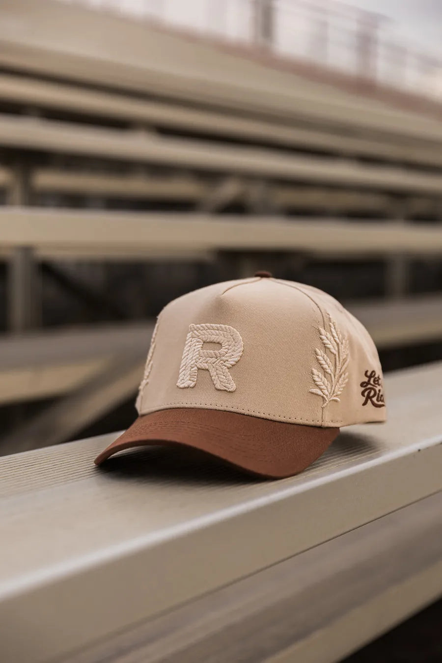 Tan rope-R hat with Let it Ride script, resting on a bleacher rail